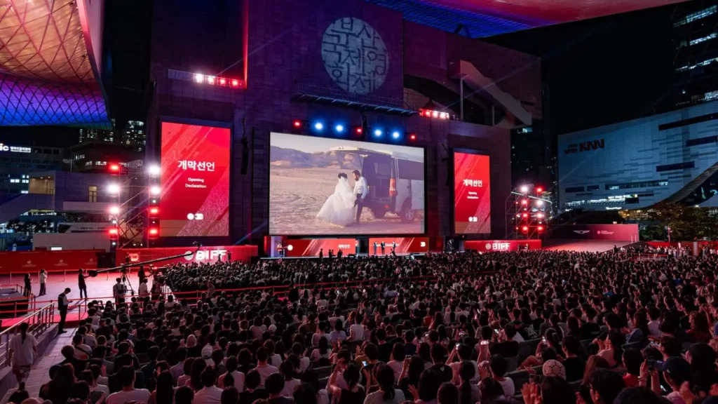 A packed outdoor audience sits facing a massive cinema screen showing a film scene at night under the illuminated canopy of the Busan Cinema Center, flanked by large vertical red screens displaying Korean and English text for the opening declaration of the Busan International Film Festival.